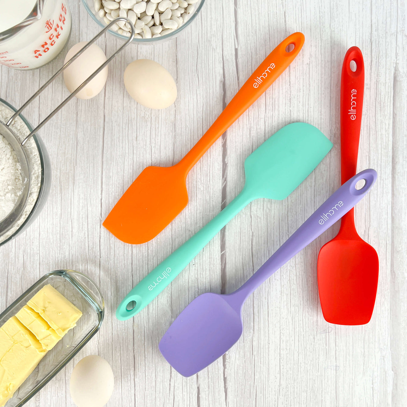 Elihome silicone spatulas in orange, mint, purple, and red displayed with baking ingredients including eggs, butter, flour, and measuring cup on white wood background