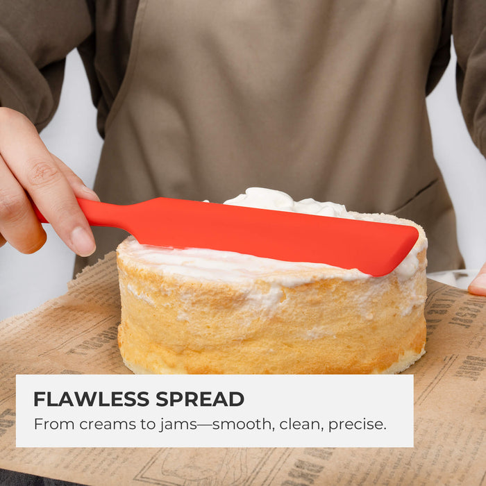 Close-up of an Elihome red skinny silicone spatula spreading cream frosting on a cake layer to demonstrate control. Text reads: "Flawless Spread. From creams to jams—smooth, clean, precise."