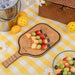 Fruit salad on a paddle-shaped cutting board with a yellow checkered tablecloth, picnic basket, and lemon on a wooden surface.