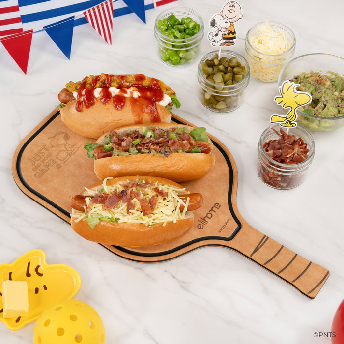 Three hot dogs on a wooden paddle board with various condiments and a Snoopy figurine in the background.