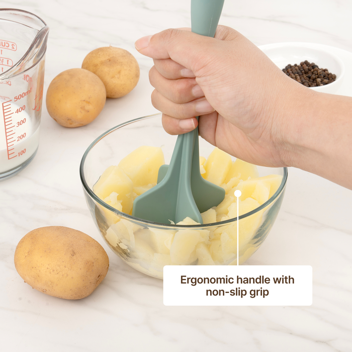 Hand mashing boiled potatoes in a glass bowl with an Elihome Classic Chopper in Sage Green, ergonomic handle with non-slip grip