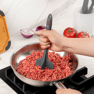 Hand using an Elihome Classic Chopper in Grey to break up ground beef in a stainless steel skillet on the stove, beside a red onion and tomatoes