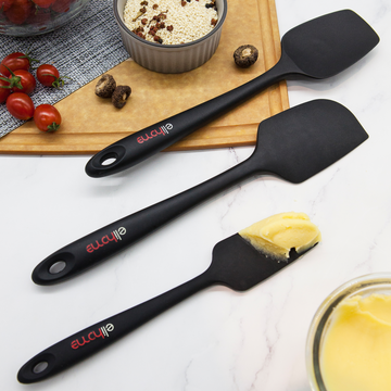 Two Elihome Mini silicone spatulas in Black on a marble counter, one lifting a pat of butter, beside cherry tomatoes and a ramekin of crumble topping