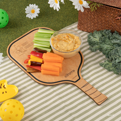 Elihome x Peanuts pickleball paddle board with hummus, celery, carrots and bell peppers at a picnic on the grass
