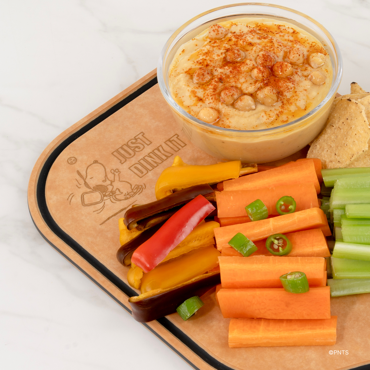 Close-up of an Elihome x Peanuts pickleball paddle board engraved with Snoopy, served with hummus and vegetable sticks