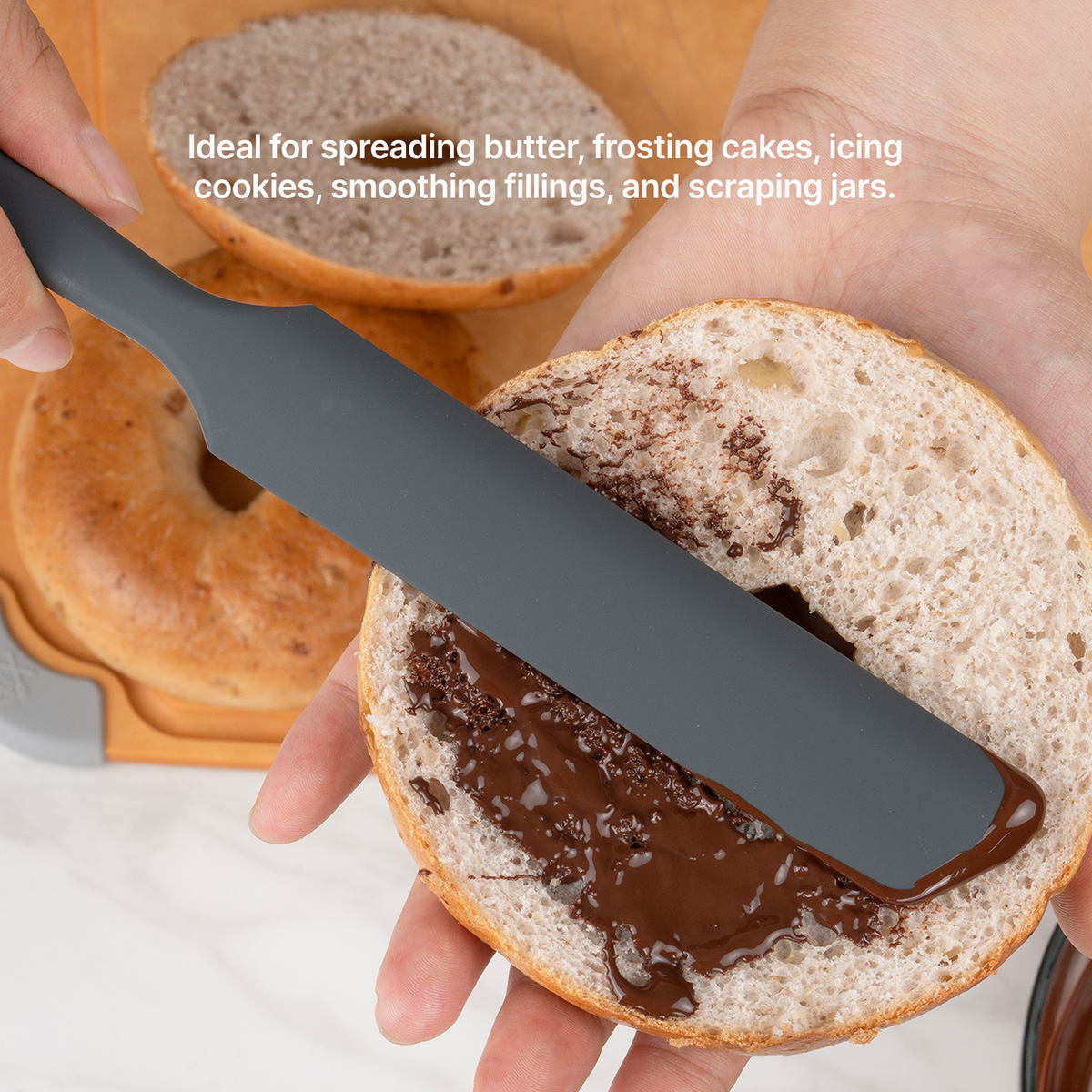 Hand using the Elihome x Peanuts Silicone Spreader to spread chocolate on a bagel half, ideal for spreading butter, frosting cakes, icing cookies, smoothing fillings and scraping jars