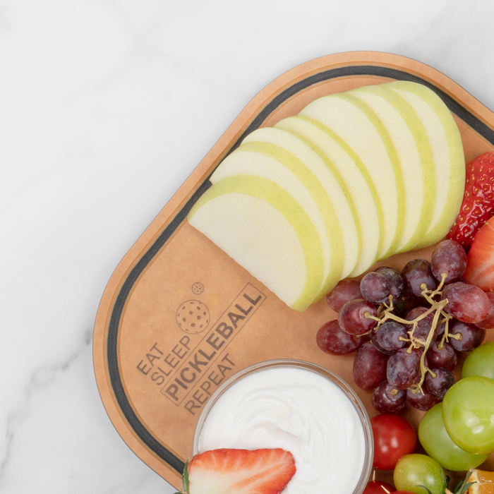Apple slices, red and green grapes, strawberries, and a bowl of yogurt dip served on an Elihome pickleball paddle board engraved with Eat Sleep Pickleball Repeat